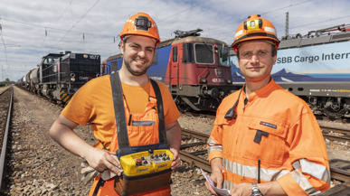 Zwei SBB-Carco-International-Mitarbeitende in stolzer Haltung und oranger Arbeitskleidung vor einer SBB-Cargo-Lok