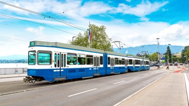 Ein altes weiss blaues Tram, das über eine Brücke fährt. Im Hintergrund sieht man einen Teil eines Sees und Teil der Stadt