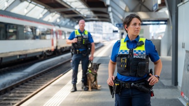 Zwei SBB Beamte stehen auf dem Perron in einem Bahnhof. Einer der Beamten ist näher an der Kamera und ist weiblich, der hintere ist männlich und hat einen Deutschenschäferhund neben sich