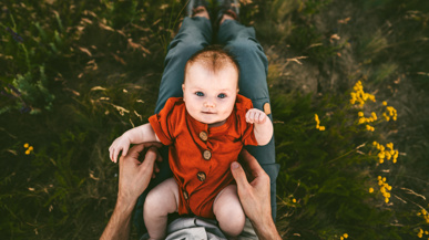 Ein Bild eines Babys in rotem Anzug, das auf dem Schoss des Vaters liegt und nach oben schaut