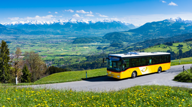 Ein Postauto auf einer Landstrasse mit grossen Wiesen und Bergen im Hintergrund