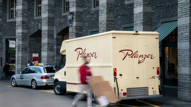 Ein Mitarbeiter von Planzer mit rotem Pullover liefert Pakete aus; hinter ihm ein Planzer-Lastwagen mit rotem Schriftzug auf weiss