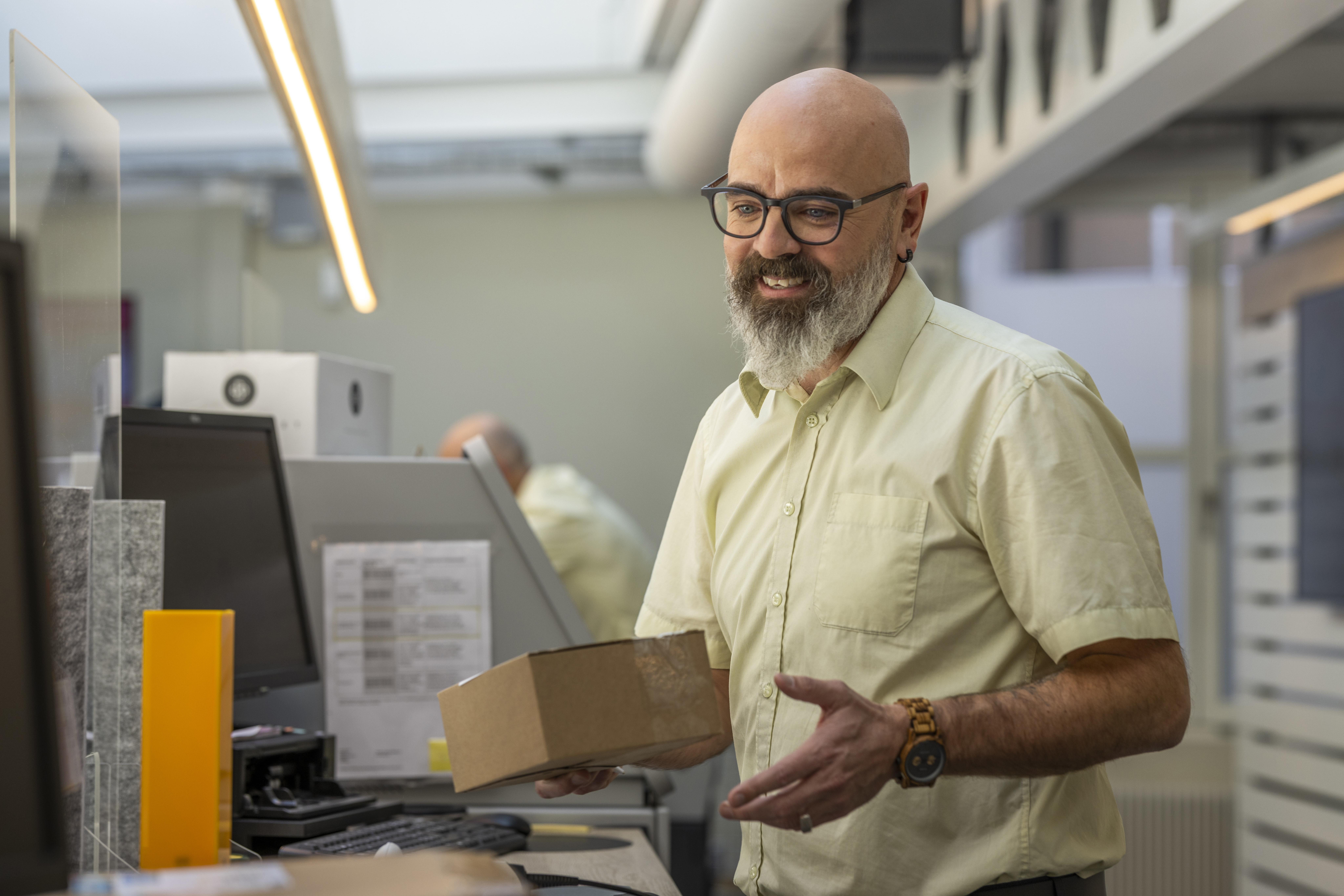 Ein Postbeamter mit Glatze, Brille und gelbem Hemd arbeitet am Schalter und hat ein Paket in der Hand